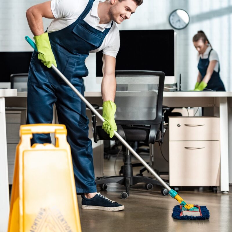 smiling-cleaner-washing-floor-with-mop-near-colleague-cleaning-desk.jpg